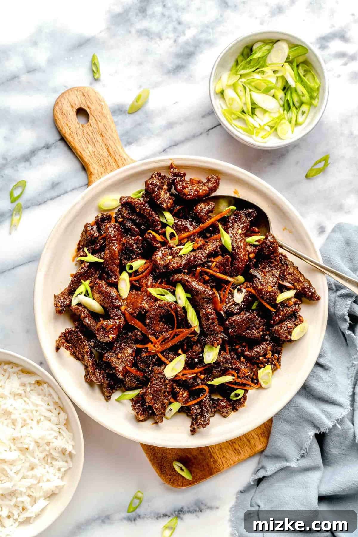 Crispy beef ready to be garnished and enjoyed with rice. An overhead shot of crispy beef on a serving platter, with a bowl of white rice and a small bowl of chopped scallions nearby.