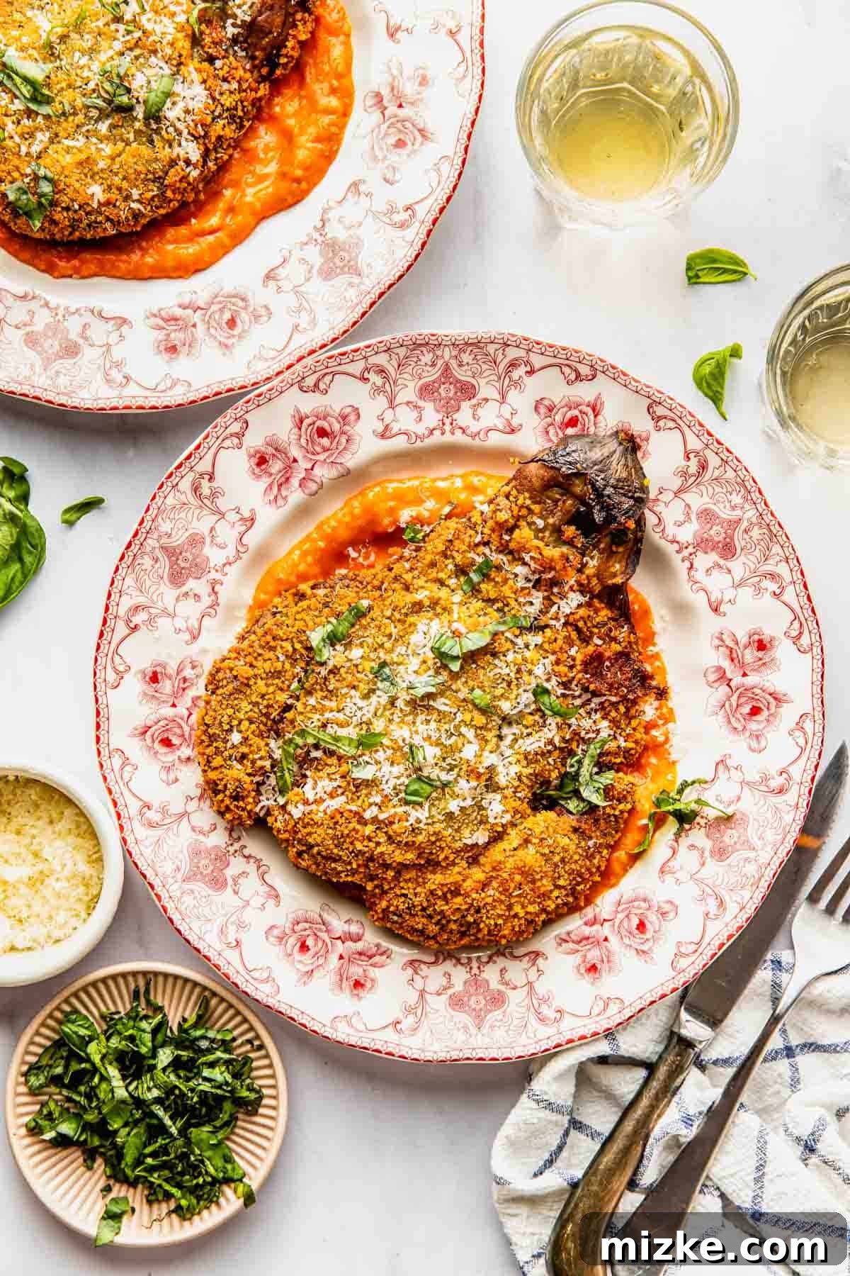 Crispy Fried Eggplant Cutlets 2 A beautifully breaded and fried eggplant schnitzel rests on a vibrant red-and-white plate, generously coated in a luscious tomato sauce and garnished with fresh herbs and grated Parmesan cheese. Accompanying the dish are small bowls of additional cheese and finely chopped herbs, alongside elegant glasses of white wine, suggesting a perfect meal setting.