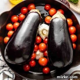 Crispy Fried Eggplant Cutlets 5 Two whole eggplants, cherry tomatoes, and a head of garlic, all drizzled with olive oil in a cast iron skillet, ready for roasting. Salt, pepper, and fresh basil are seen nearby.