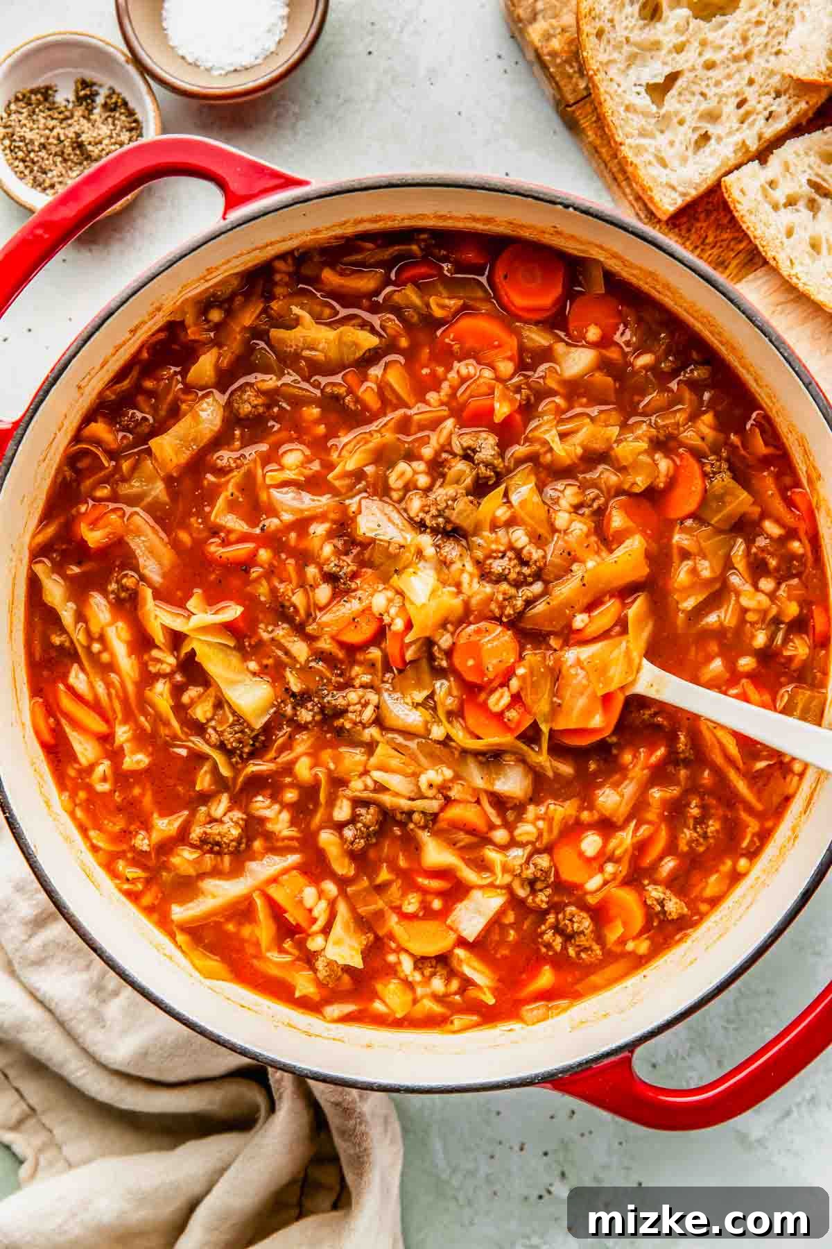 Hearty Beef Barley and Cabbage Soup 3 A large pot of simmering beef and cabbage soup, filled with rich tomato broth, ground beef, carrots, and softened cabbage, on a kitchen counter next to serving utensils and fresh bread.