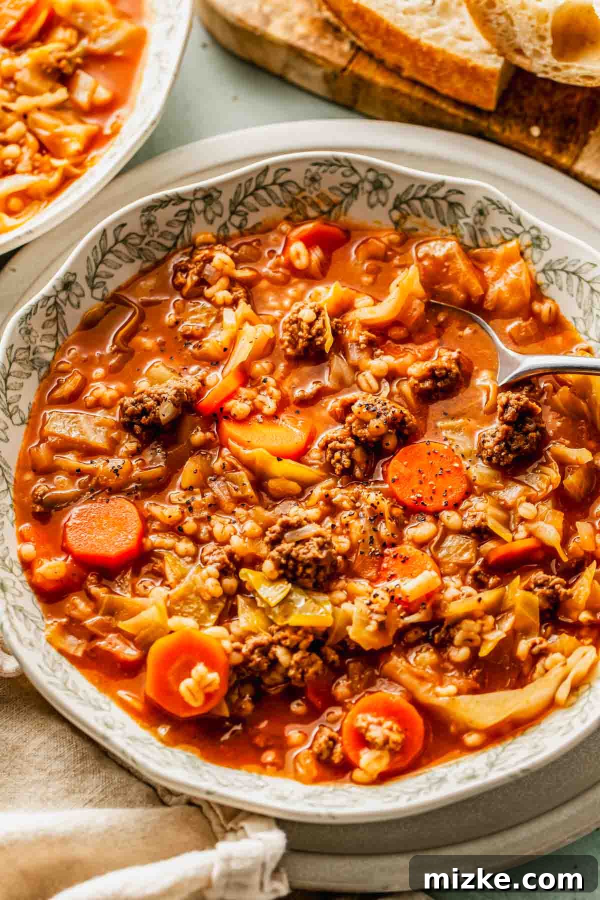Hearty Beef Barley and Cabbage Soup 4 A close-up of a serving of beef barley and cabbage soup, showcasing the chunks of ground beef, visible barley grains, sliced carrots, and tender cabbage in a rich, red broth, with a spoon ready for tasting.