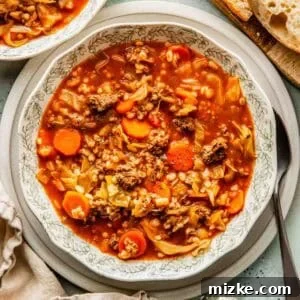 A bowl of beef and barley soup with carrots, cabbage, ground meat, and barley, served with slices of bread on the side.