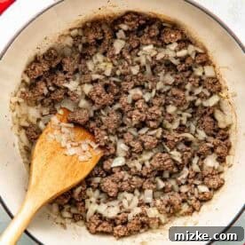 Ground beef and chopped onions are being cooked in a large pot, with a wooden spoon resting inside.