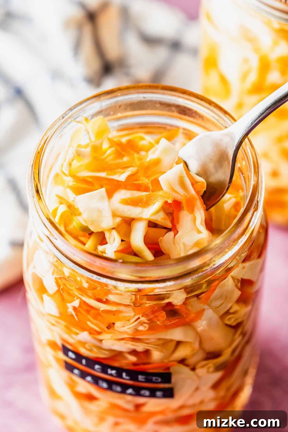 A clear glass jar containing vibrant, finely shredded pickled cabbage and carrots, with a rustic striped cloth draped in the background. Another partially visible jar adds to the artisanal feel, suggesting a fresh, homemade batch.