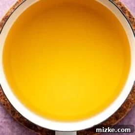 A white saucepan filled with clear yellow brining liquid, placed on a round cork pad against a purple textured background.