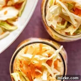 Two glass jars filled with shredded cabbage and carrots, with more of the vegetable mixture visible on a plate in the background.