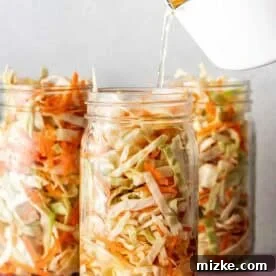 Three glass jars filled with shredded cabbage and carrots, with liquid being poured into one jar from a white container.