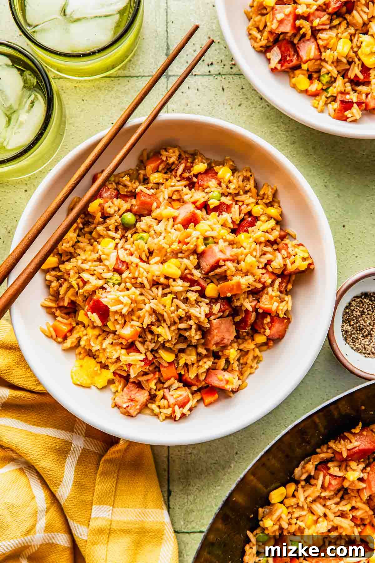 Crispy Garlic Spam Fried Rice with Vegetables 3 A bowl of fried rice with vegetables and diced meat, served with chopsticks, ice water, and a small bowl of pepper on a light green surface.