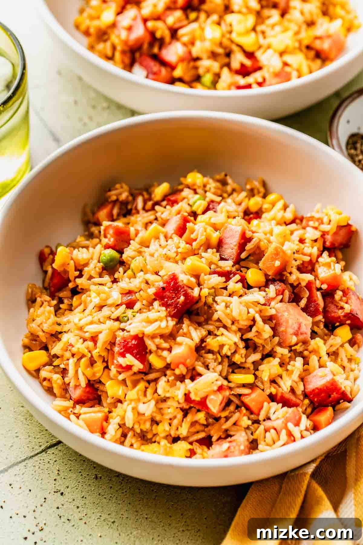 Crispy Garlic Spam Fried Rice with Vegetables 4 A white bowl filled with fried rice mixed with diced ham, corn, carrots, and peas. A second bowl of the same dish is partially visible in the background.