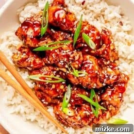 general tso's chicken on top of white rice in a bowl with wooden chopsticks. garnished with sesame seeds and scallions
