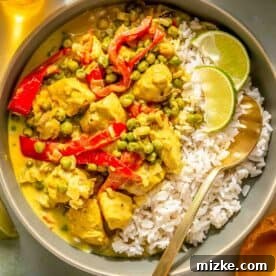 Bowl of green curry with chicken, red peppers, peas, and rice, garnished with lime wedges.