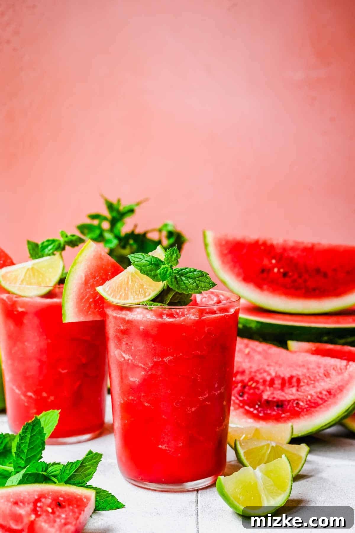 Two glasses of watermelon slushies garnished with lime wedges and mint, surrounded by watermelon slices, lime pieces, and mint leaves on a white surface against a pink background.