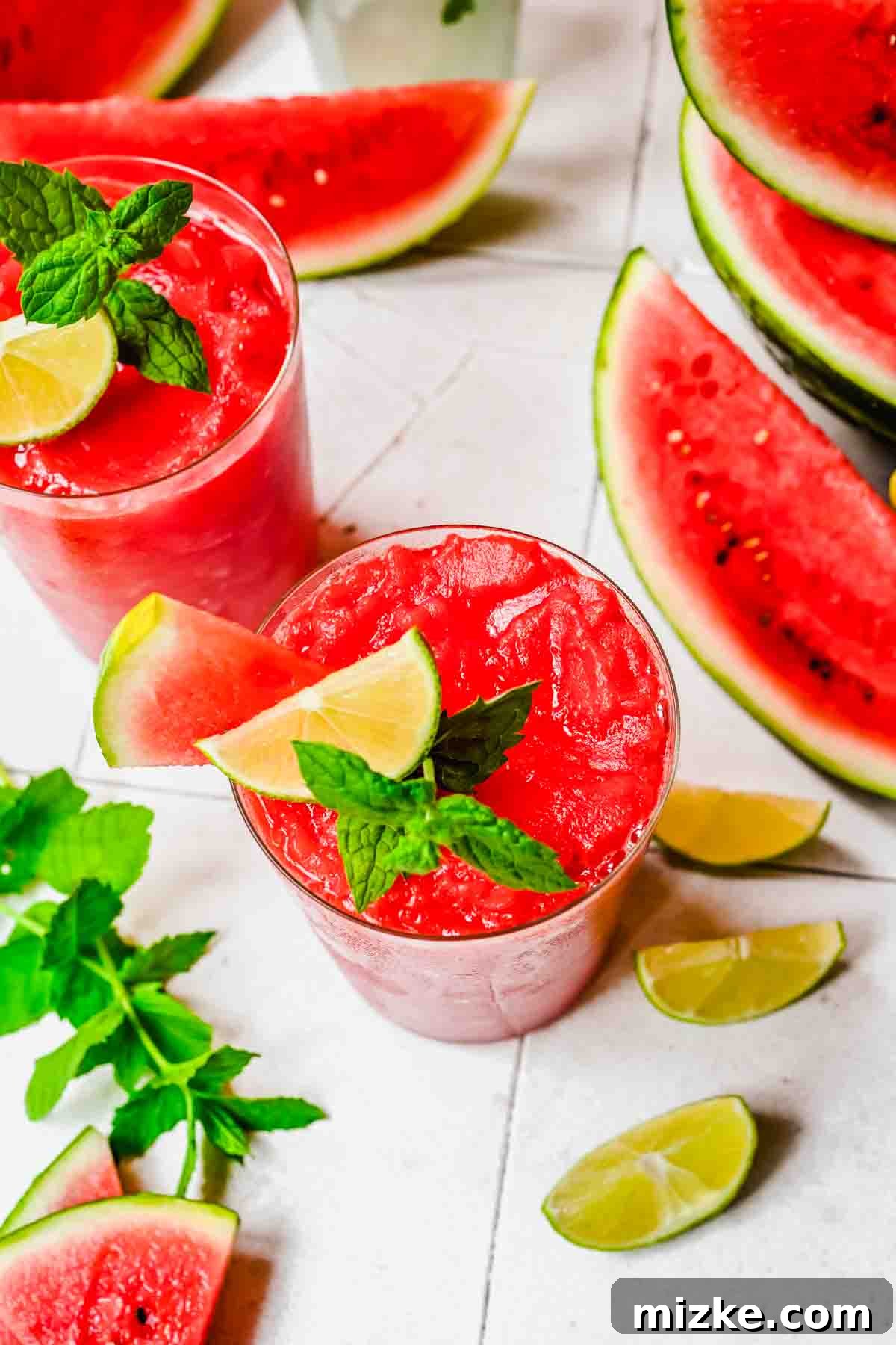 Two glasses of watermelon slushies garnished with lime, mint, and watermelon slice, surrounded by watermelon wedges, lime wedges, and mint leaves on a white surface.