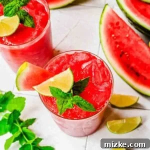 Two glasses of watermelon slushies garnished with lime, mint, and watermelon slice, surrounded by watermelon wedges, lime wedges, and mint leaves on a white surface.