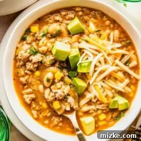 A bowl of white chili with ground turkey, corn, beans, shredded cheese, avocado cubes, and herbs, served with a spoon.