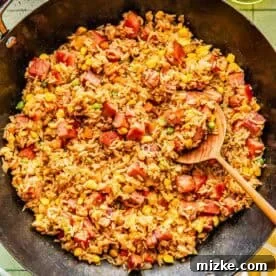 A skillet filled with fried rice mixed with diced ham, corn, peas, and carrots, with a wooden spoon resting inside.