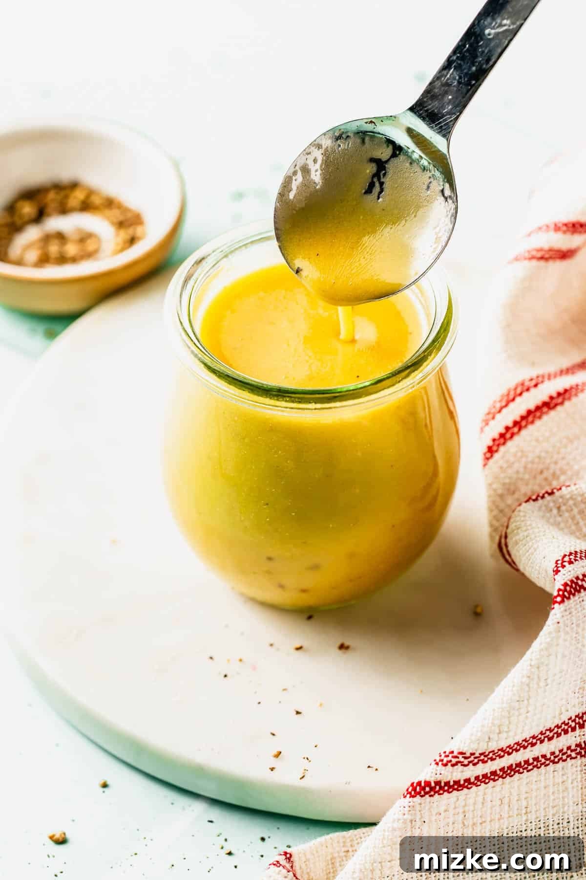 Apple Cider Vinaigrette Recipe for Fresh Salads 2 A spoon is held above a small glass jar filled with yellow salad dressing; a red-striped cloth and a dish of seasoning sit nearby.