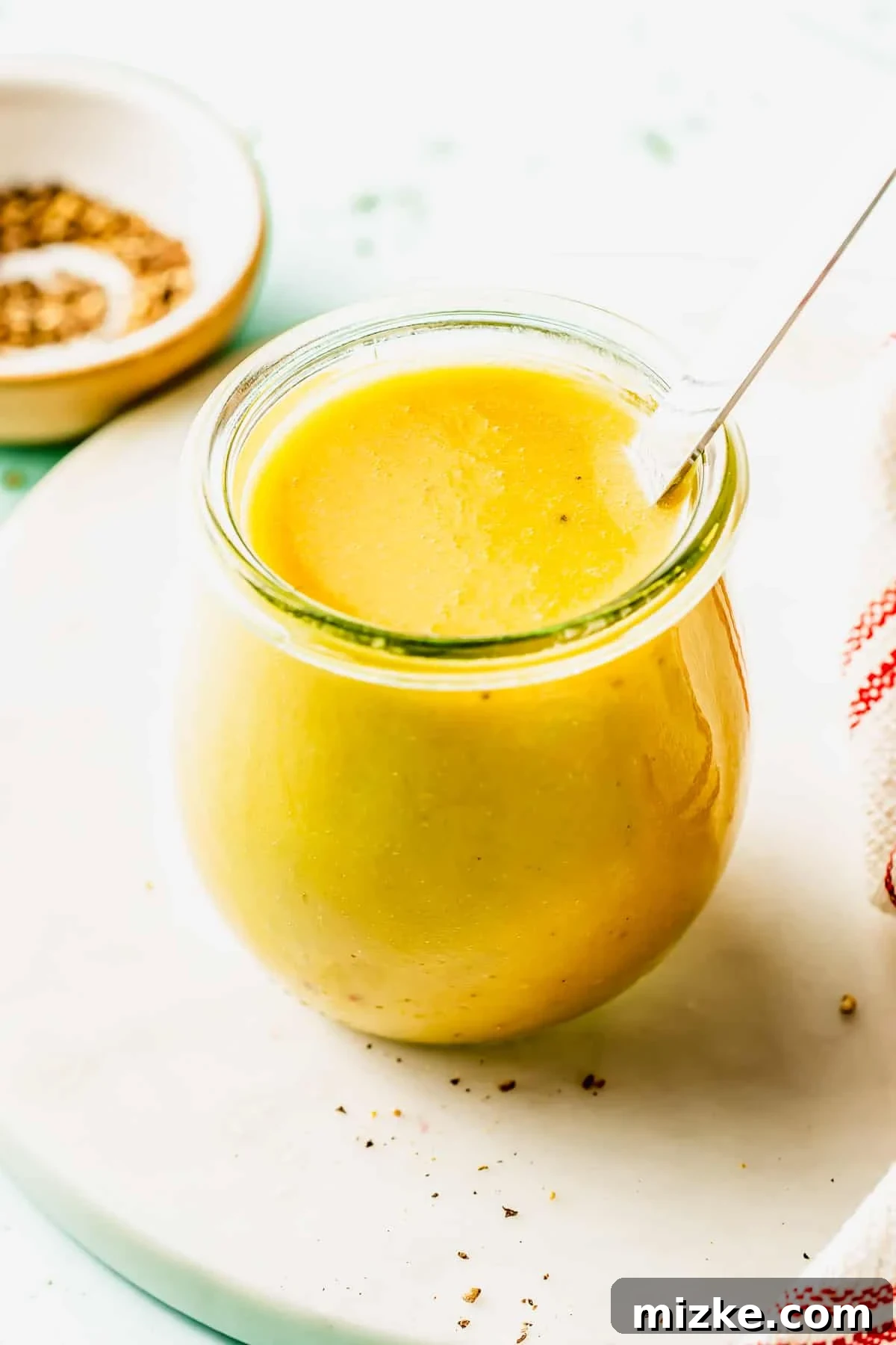 Apple Cider Vinaigrette Recipe for Fresh Salads 3 A glass jar filled with yellow liquid, possibly salad dressing or sauce, with a spoon inside. A small dish with spices and a striped cloth are nearby on a white surface.