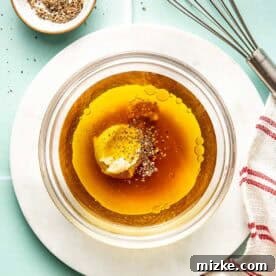 Apple Cider Vinaigrette Recipe for Fresh Salads 5 A glass bowl with oil, mustard, and pepper, a small bowl of pepper, a metal whisk, and a red-striped towel on a light surface.