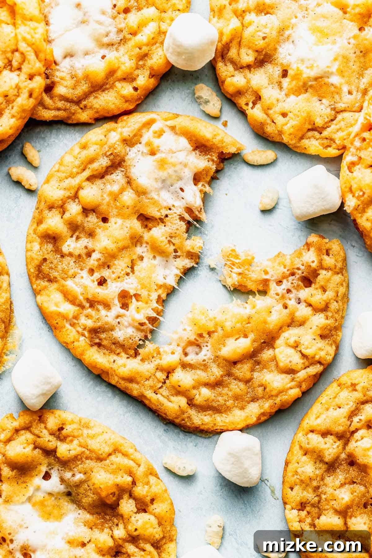 Crispy Marshmallow Cookies with Gooey Centers 4 A close-up of crispy cookies with bits of marshmallow and rice cereal, one cookie partially eaten, surrounded by small marshmallows.